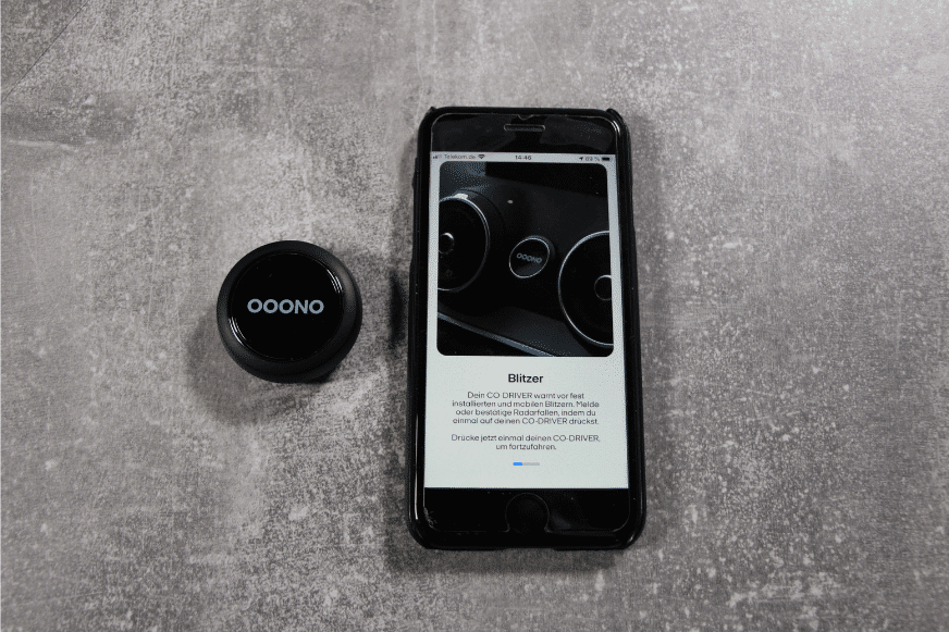 Der Ooono CO-DRIVER NO1 liegt neben dem Smartphone und wird für eine Blitzerwarnung 1 Mal gedrückt.